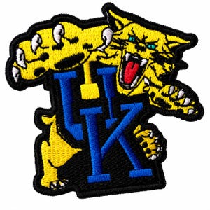 May include: An embroidered patch featuring a yellow wildcat with black spots, white claws, and a red tongue, positioned above and around the blue letters UK. The design is outlined in black thread with a solid black background border.