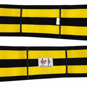 May include: A rectangular fitness belt featuring horizontal black and yellow stripes. The belt includes a white fabric patch in the center displaying the red and black text 'dk' above the words 'DRAGON FITNESS' and a small white care label tag.