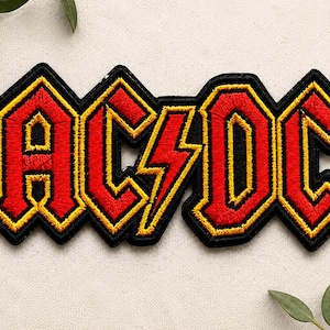 May include: An embroidered fabric patch featuring the text AC/DC in a bold, stylized font. The letters are bright red with a thick yellow border, all outlined in black. A yellow lightning bolt symbol is positioned between the letters C and D.