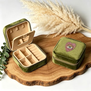 May include: Two square olive green velvet jewelry boxes with beige interiors. One box is open, revealing a mirror, ring rolls, and compartments. The other box is closed and features an embroidered patch of a heart with an eye on the lid.