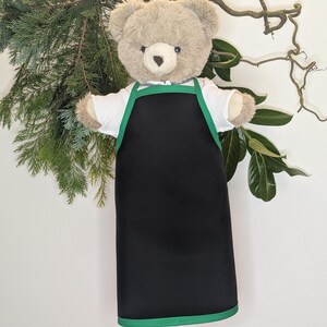 May include: A plush teddy bear wearing a black apron with green trim over a white shirt. The bear has a light brown fur and is positioned in front of a backdrop of green foliage and branches.