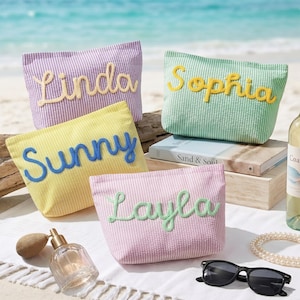 Op de afbeelding: Vier pastelkleurige seersucker cosmetische tassen, elk met een naam geborduurd in een contrasterende kleur: "Linda", "Sophia", "Sunny" en "Layla". De tassen zijn in lavendel, mintgroen, geel en roze. De afbeelding bevat zonnebrillen, parfum en een strand.