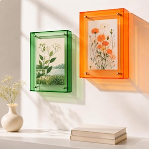 May include: Two rectangular acrylic wall frames, one in translucent green and one in translucent orange, each secured with four silver-toned metal corner bolts. The green frame displays a botanical print of white flowers, while the orange frame holds a print of orange and white cosmos flowers.