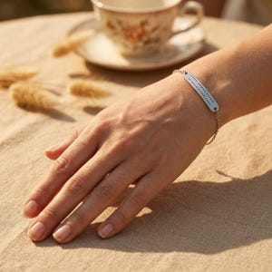 Könnte beinhalten: Ein silbernes Armband mit einem rechteckigen Steg und einer feinen Kette. Der Steg weist eine Reihe kleiner, gleichmäßig verteilter Punkte auf. Das Armband wird am Handgelenk getragen, mit einer Tasse und Untertasse im Hintergrund.