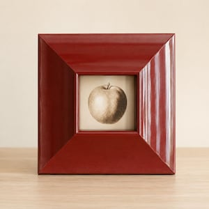 Puede incluir: Un marco de fotos cuadrado con un acabado rojo oscuro brillante y bordes anchos e inclinados. El marco contiene una impresión cuadrada centrada que presenta un dibujo detallado al carboncillo monocromático de una manzana.