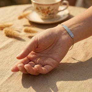 May include: A silver bracelet with a rectangular tag and a delicate chain is worn on a wrist. The tag has small, evenly spaced markings. The background includes a teacup and saucer, and dried plants on a tan surface.