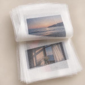 May include: A stack of translucent white tissue paper sheets, each featuring a rectangular printed image. The top sheet displays a sunset over the ocean, while the sheet beneath shows a window view of the sea with curtains and a vase of flowers.