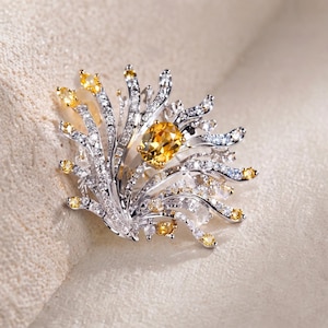 May include: A silver-toned brooch featuring a central oval-cut yellow citrine gemstone surrounded by radiating, curved metal branches. The branches are encrusted with numerous small, clear white stones and accented with smaller yellow stones.