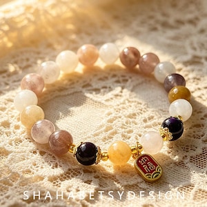 Pode incluir: Uma pulseira de contas com pedras redondas e lisas em tons de branco, pêssego, castanho, amarelo e roxo escuro. A pulseira inclui espaçadores de metal dourado e um pequeno pendente retangular vermelho com caracteres chineses dourados.