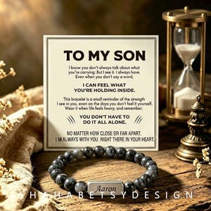 Puede incluir: Una pulsera de cuentas de piedra gris oscuro con una barra metálica rectangular plateada en el centro grabada con el nombre Aaron. La pulsera se sitúa frente a una tarjeta cuadrada con el texto impreso TO MY SON y un mensaje.
