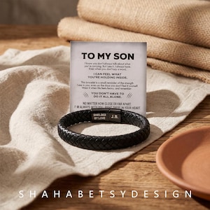 May include: A black braided leather bracelet with a rectangular metal clasp featuring the engraved text SHIELDED BY LOVE and J.B. The bracelet is displayed in front of a white card with the printed text TO MY SON and a sentimental message.