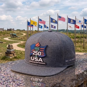 May include: A heather grey baseball cap with a flat brim and a patch on the front featuring a torch, the number 250, the text USA, and the word SEMIQUINCENTENNIAL. The side of the cap features the text WE THE PEOPLE in white lettering.
