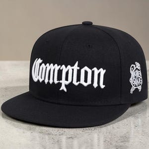 May include: Black baseball cap with the word "Compton" embroidered in white script across the front. The cap features a flat brim and a small embroidered design on the side, with a button on top.