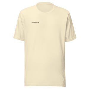 以下が含まれることがあります： クリーム色の半袖Tシャツ。テキスト「not finished yet」は、胸の左上に小さな黒い文字で印刷されています。シャツはクルーネックで、ゆったりとしたフィット感です。