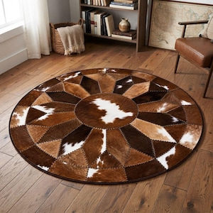 May include: A circular area rug constructed from stitched panels of cowhide in varying shades of brown, tan, and white. The rug features a geometric starburst pattern radiating from a central circular patch, with visible stitching along each seam.