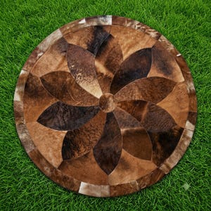May include: A circular rug constructed from individual pieces of cowhide leather arranged in a radial, petal-like pattern. The hide segments feature a natural color palette of brown, tan, dark chocolate, and cream tones with varied textures.
