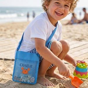 Può includere: Una borsa a tracolla in rete azzurra con il nome Charlie stampato in blu scuro sopra un disegno di un granchio arancione. La borsa è riempita con piccoli sassi grigi ed è accompagnata da una paletta di plastica arancione e un gioco da sabbia.