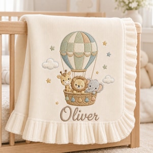 Puede incluir: Una manta de punto color crema con un bordado de un globo aerostático que transporta una jirafa, un león y un elefante. El diseño incluye pequeñas nubes y estrellas, con el nombre Oliver bordado en letras cursivas marrones debajo.