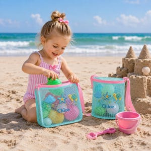 Może przedstawiać: Małe dziecko bawiące się na piaszczystej plaży z dwiema niebieskimi torbami plażowymi z siatki, r&oacute;żowym wiaderkiem i r&oacute;żową łopatką. Torby mają r&oacute;żowe wykończenia i nadrukowane imię "Chloe". Torby są wypełnione kolorowymi piłkami i zabawkami. W tle zamek z piasku.