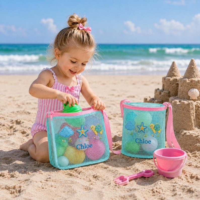 Może przedstawiać: Małe dziecko bawiące się na piaszczystej plaży z dwiema niebieskimi torbami plażowymi z siatki, r&oacute;żowym wiaderkiem i r&oacute;żową łopatką. Torby mają r&oacute;żowe wykończenia i nadrukowane imię "Chloe". Torby są wypełnione kolorowymi piłkami i zabawkami. W tle zamek z piasku.