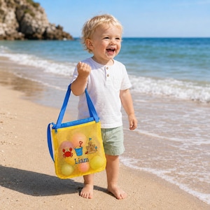 Może przedstawiać: Ż&oacute;łta torba plażowa z siatki z niebieskimi paskami, wypełniona kolorowymi piłkami, trzymana przez dziecko na piaszczystej plaży. Torba posiada grafikę kraba, zamku z piasku i wiadra, z nadrukowanym imieniem "Liam".