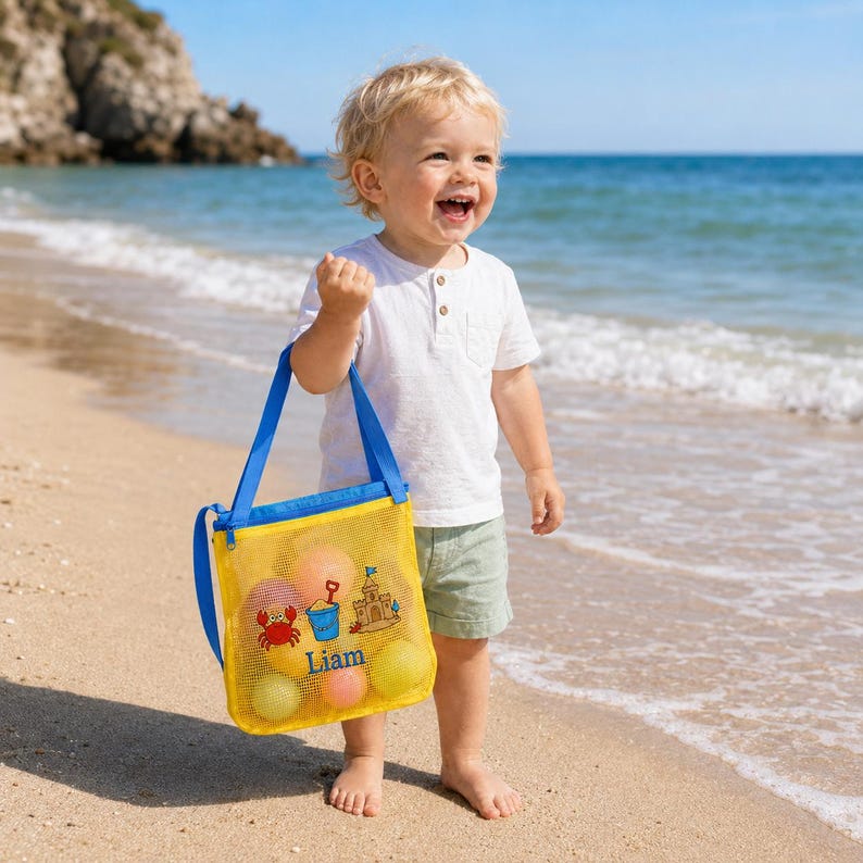 Może przedstawiać: Ż&oacute;łta torba plażowa z siatki z niebieskimi paskami, wypełniona kolorowymi piłkami, trzymana przez dziecko na piaszczystej plaży. Torba posiada grafikę kraba, zamku z piasku i wiadra, z nadrukowanym imieniem "Liam".