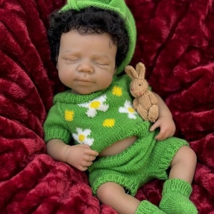 Puede incluir: Una muñeca bebé realista vestida con un conjunto de punto verde con detalles de flores blancas y amarillas, un gorro y calcetines a juego. La muñeca sostiene un pequeño conejito de punto marrón. La muñeca está sobre una manta de felpa roja.