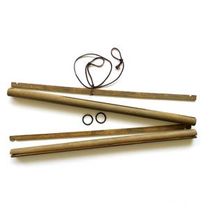 Puede incluir: Un juego de colgadores de póster de madera, con cuatro barras de madera de diferentes longitudes, una cuerda marrón y dos pequeñas gomas negras. La madera tiene un acabado natural de color marrón claro. Los artículos están dispuestos sobre un fondo blanco.