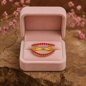 Pode incluir: Um anel protetor em ouro com duas bandas curvas ligadas nas laterais, cada uma cravejada com uma fila de pequenas pedras cor-de-rosa vibrantes. O anel &eacute; exibido dentro de uma caixa de joias de veludo rosa claro aberta e almofadada.