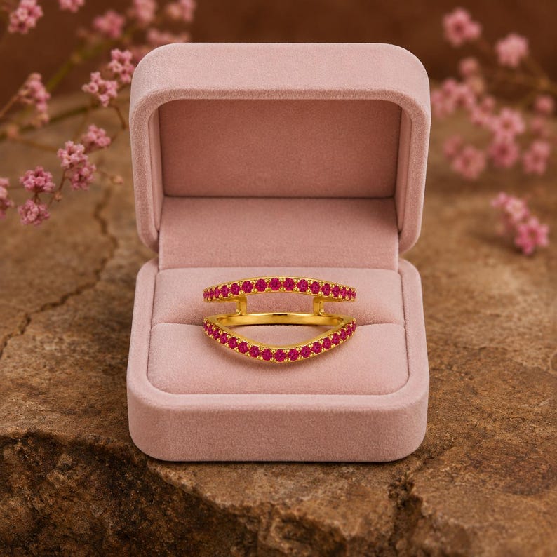 Pode incluir: Um anel protetor em ouro com duas bandas curvas ligadas nas laterais, cada uma cravejada com uma fila de pequenas pedras cor-de-rosa vibrantes. O anel &eacute; exibido dentro de uma caixa de joias de veludo rosa claro aberta e almofadada.