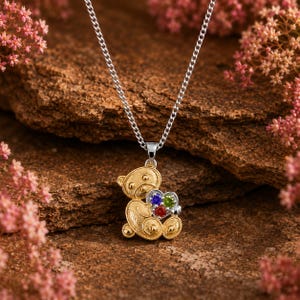 Peut inclure: Un collier avec pendentif en forme d'ourson doré présentant une finition texturée et un accent en forme de cœur argenté. Le cœur est incrusté de trois petites pierres précieuses rondes de couleur bleue, verte et rouge sur une chaîne argentée.