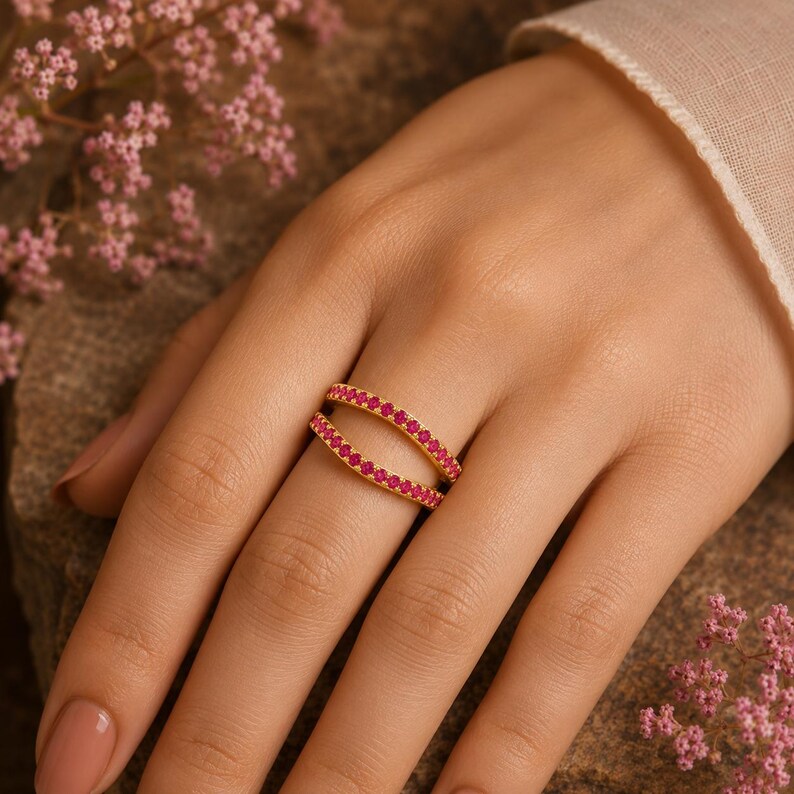 Pode incluir: Um anel de ouro com duas bandas paralelas, cada uma cravejada com uma fila cont&iacute;nua de pequenas pedras cor-de-rosa vibrantes. O design cria uma silhueta aberta e curva que envolve o dedo, exibindo um acabamento met&aacute;lico polido.