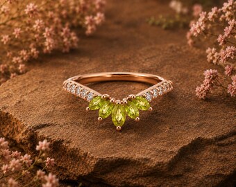 May include: A rose gold ring with a curved band, set with small white diamonds and five pear-shaped, light green gemstones. The ring is displayed on a brown stone surface, with pink flowers in the background.