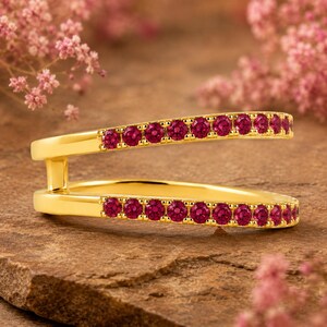 Pode incluir: Um anel aberto de ouro com duas bandas paralelas ligadas por uma barra vertical. Cada banda apresenta uma fila de pequenas pedras preciosas redondas de um vermelho rosado vibrante, dispostas nas sec&ccedil;&otilde;es superior e inferior do anel.