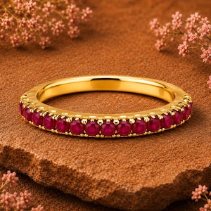 Op de afbeelding: Een gouden ring met een band van kleine, ronde, dieprode edelstenen. De ring ligt op een getextureerd, bruin oppervlak, met roze bloemen op de achtergrond. De gouden band van de ring heeft een getextureerd ontwerp.