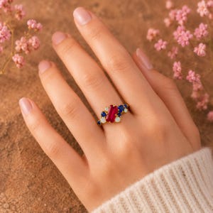 Op de afbeelding: Een gouden ring met een centrale rechthoekige rode edelsteen, geflankeerd door clusters van kleinere ronde blauwe saffieren en witte opalen. De stenen zijn gezet in een geelgouden band, wat zorgt voor een meerstenig ontwerp.