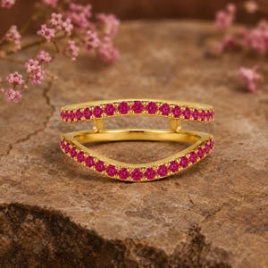 May include: A gold ring featuring a double-band design with an open, curved silhouette. Both bands are encrusted with small, vibrant pink circular gemstones set in a continuous row, creating a bright and colorful appearance on the metal frame.