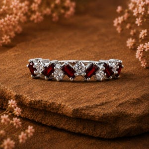 Pode incluir: Um anel de prata com pedras preciosas vermelhas e claras alternadas. O anel apresenta pedras vermelhas retangulares e pedras claras redondas. O anel está colocado sobre uma superfície marrom com flores rosas ao fundo.