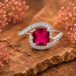 Può includere: Un anello color argento con una grande pietra preziosa rosso vivo a taglio cuscino al centro. La fascia presenta una forma asimmetrica a spirale, incastonata con piccole pietre chiare e scintillanti su tutta la superficie.