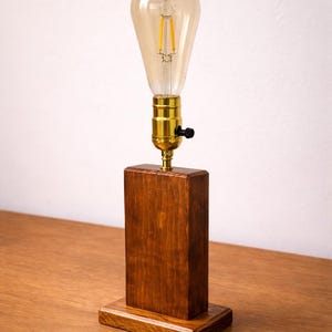 Puede incluir: Una lámpara de mesa de madera con un acabado marrón cálido. La lámpara presenta una base rectangular y un casquillo de bombilla de color latón. Una gran bombilla en forma de lágrima está atornillada en el casquillo. La lámpara mide unos 30 cm de alto.