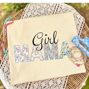 Op de afbeelding: Een lichtgeel t-shirt met de tekst "Girl Mama". Het woord "Girl" is in zwart script, terwijl "Mama" in een bloemenpatroon met een lichtblauwe omtrek staat. Het shirt ligt op een rieten mand.