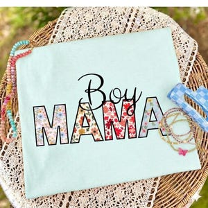 Op de afbeelding: Lichtblauw t-shirt met de tekst "Boy Mama" in zwart script en letters met bloemenprint. Het shirt ligt op een gevlochten mand met kralenarmbanden en een blauwe zonnebril.