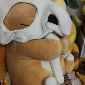 K&ouml;nnte beinhalten: Ein braunes Cubone-Pok&eacute;mon-Pl&uuml;schtier mit einer wei&szlig;en Totenkopfmaske. Das Spielzeug hat einen gro&szlig;en Kopf und einen kleinen K&ouml;rper. Im Hintergrund sind weitere Pok&eacute;mon-Pl&uuml;schtiere zu sehen, darunter ein gr&uuml;ner, drachenartiger Charakter.