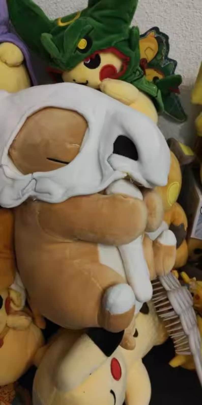 K&ouml;nnte beinhalten: Ein braunes Cubone-Pok&eacute;mon-Pl&uuml;schtier mit einer wei&szlig;en Totenkopfmaske. Das Spielzeug hat einen gro&szlig;en Kopf und einen kleinen K&ouml;rper. Im Hintergrund sind weitere Pok&eacute;mon-Pl&uuml;schtiere zu sehen, darunter ein gr&uuml;ner, drachenartiger Charakter.