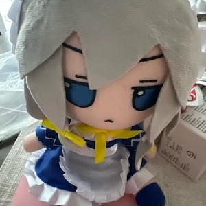 May include: A plush doll featuring a character with gray hair, blue eyes, and a blue and white dress with a yellow bow. The doll has a stern expression and is made of soft fabric.