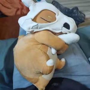 K&ouml;nnte beinhalten: Ein Cubone Pok&eacute;mon-Pl&uuml;schtier mit einem braunen K&ouml;rper und einem wei&szlig;en Sch&auml;delhelm. Das Spielzeug liegt mit verschr&auml;nkten Armen. Der Sch&auml;del hat schwarze Augenh&ouml;hlen und eine kleine Nase. Das Spielzeug liegt auf einer blauen Oberfl&auml;che.