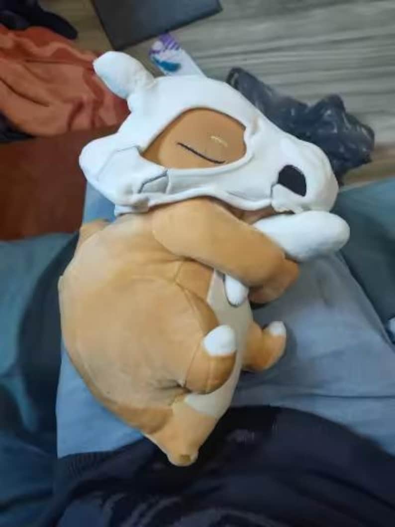 K&ouml;nnte beinhalten: Ein Cubone Pok&eacute;mon-Pl&uuml;schtier mit einem braunen K&ouml;rper und einem wei&szlig;en Sch&auml;delhelm. Das Spielzeug liegt mit verschr&auml;nkten Armen. Der Sch&auml;del hat schwarze Augenh&ouml;hlen und eine kleine Nase. Das Spielzeug liegt auf einer blauen Oberfl&auml;che.