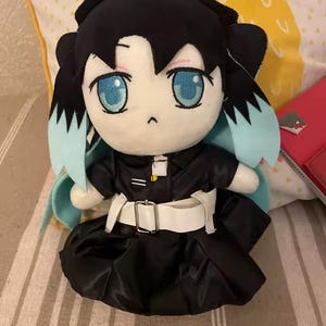 May include: A plush doll featuring black hair with light blue tips, large blue eyes, and a small mouth. The doll wears a black collared outfit with a white belt featuring a silver buckle, a white collar detail, and a small yellow accent on the chest.