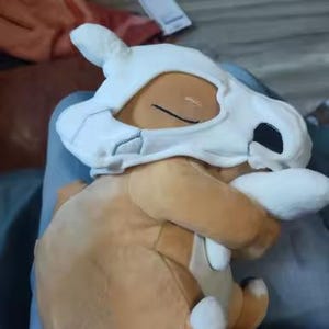 K&ouml;nnte beinhalten: Ein Cubone Pok&eacute;mon-Pl&uuml;schtier. Das Spielzeug ist haupts&auml;chlich braun mit einer wei&szlig;en Sch&auml;delmaske und wei&szlig;en Details an F&uuml;&szlig;en und H&auml;nden. Das Cubone befindet sich in einer sitzenden Position.