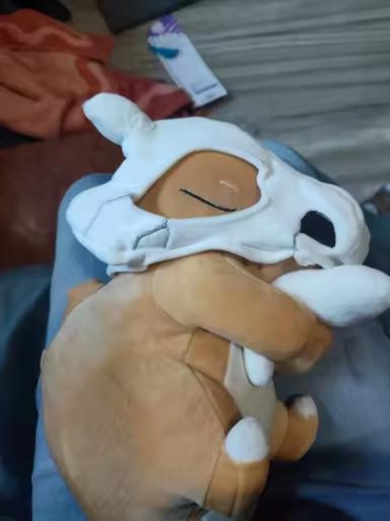 K&ouml;nnte beinhalten: Ein Cubone Pok&eacute;mon-Pl&uuml;schtier. Das Spielzeug ist haupts&auml;chlich braun mit einer wei&szlig;en Sch&auml;delmaske und wei&szlig;en Details an F&uuml;&szlig;en und H&auml;nden. Das Cubone befindet sich in einer sitzenden Position.